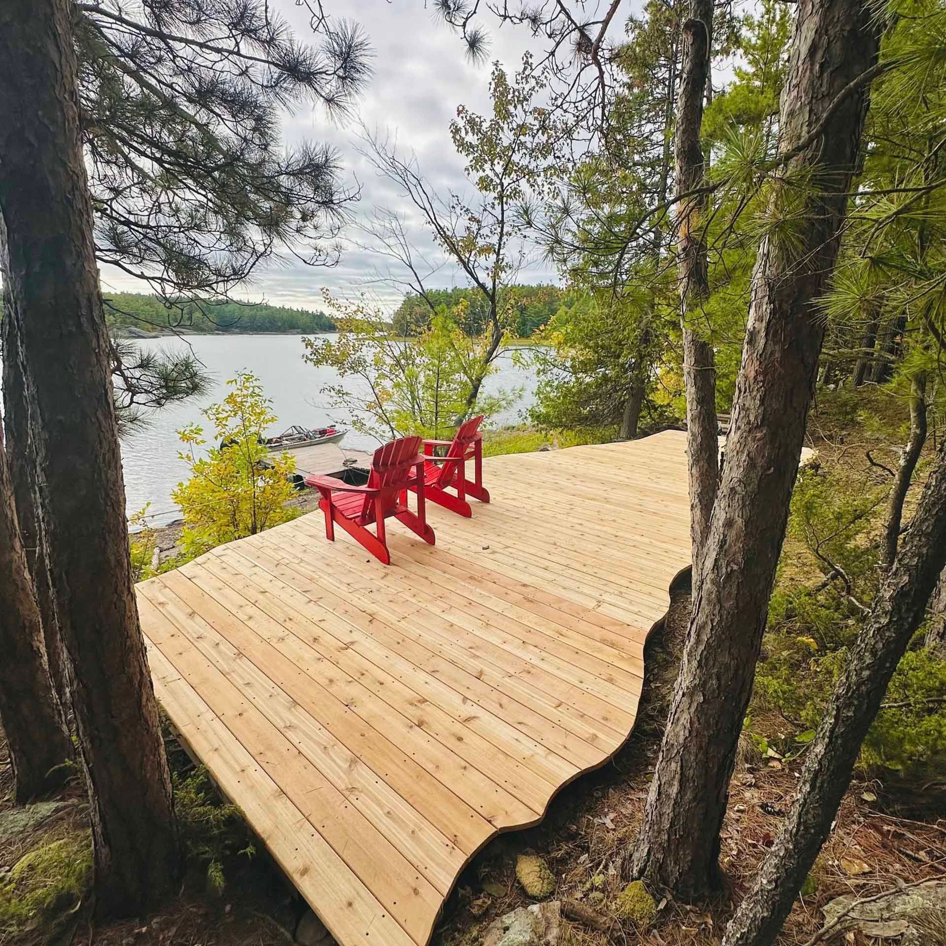 Wooden deck with two red chairs overlooking a lake. Trees frame the view.