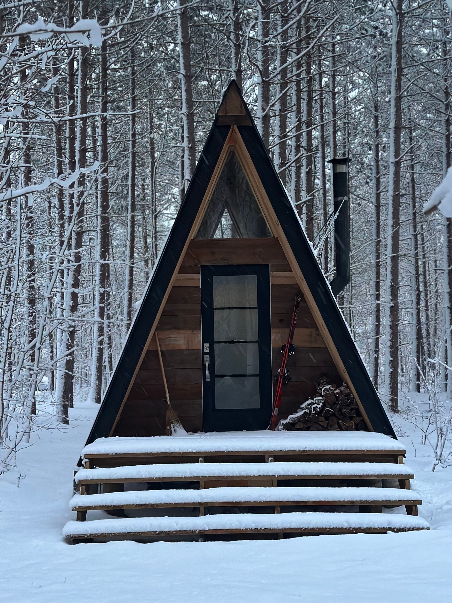 A-frame cabin in a snowy forest; black door, chimney, wooden steps, and snow-covered ground.