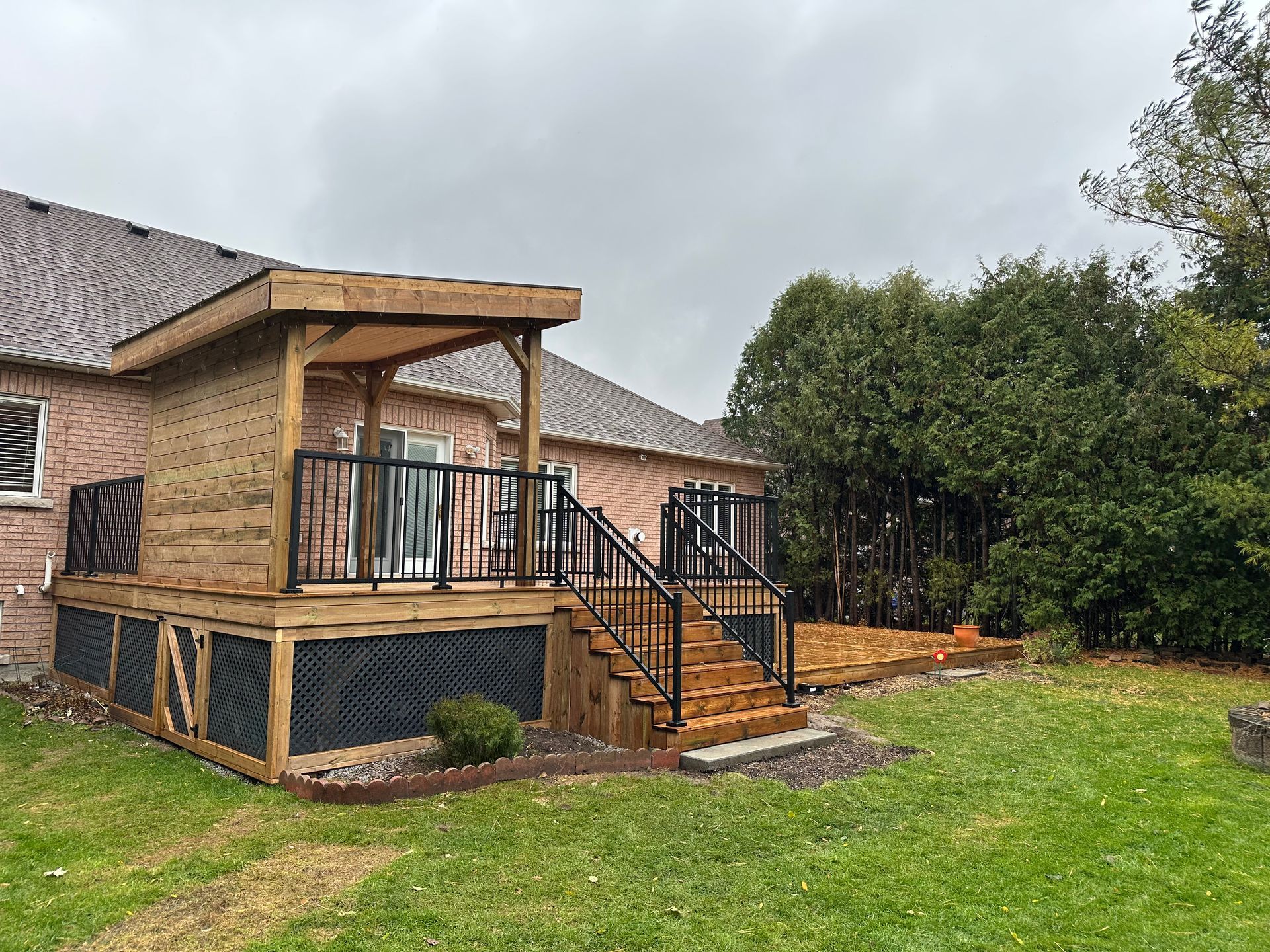 Wooden deck with black railings and steps attached to a brick house, next to a grassy yard.