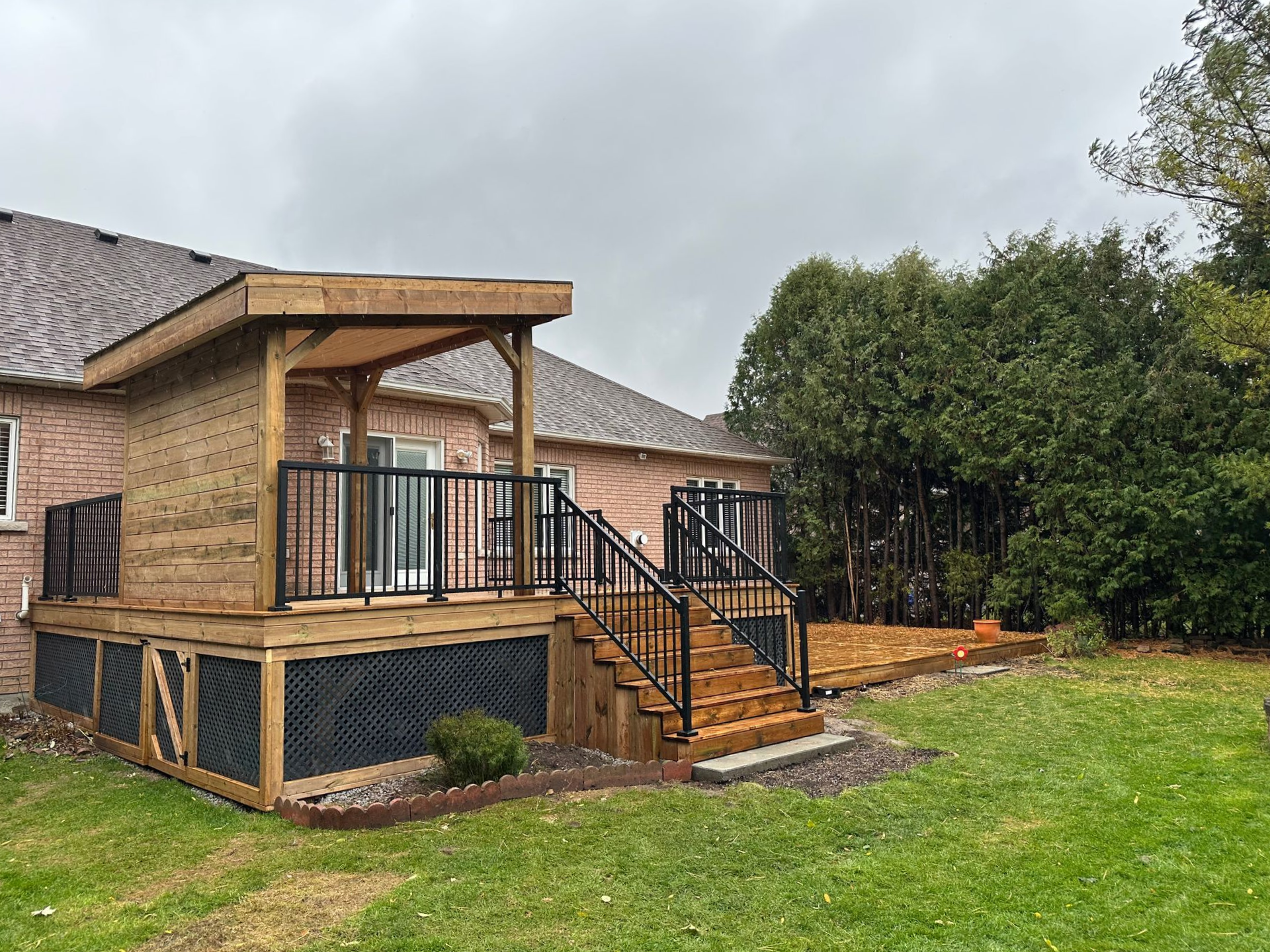 Wooden deck attached to a brick house. Stairs and black metal railing lead to a lower deck area. Overcast sky.