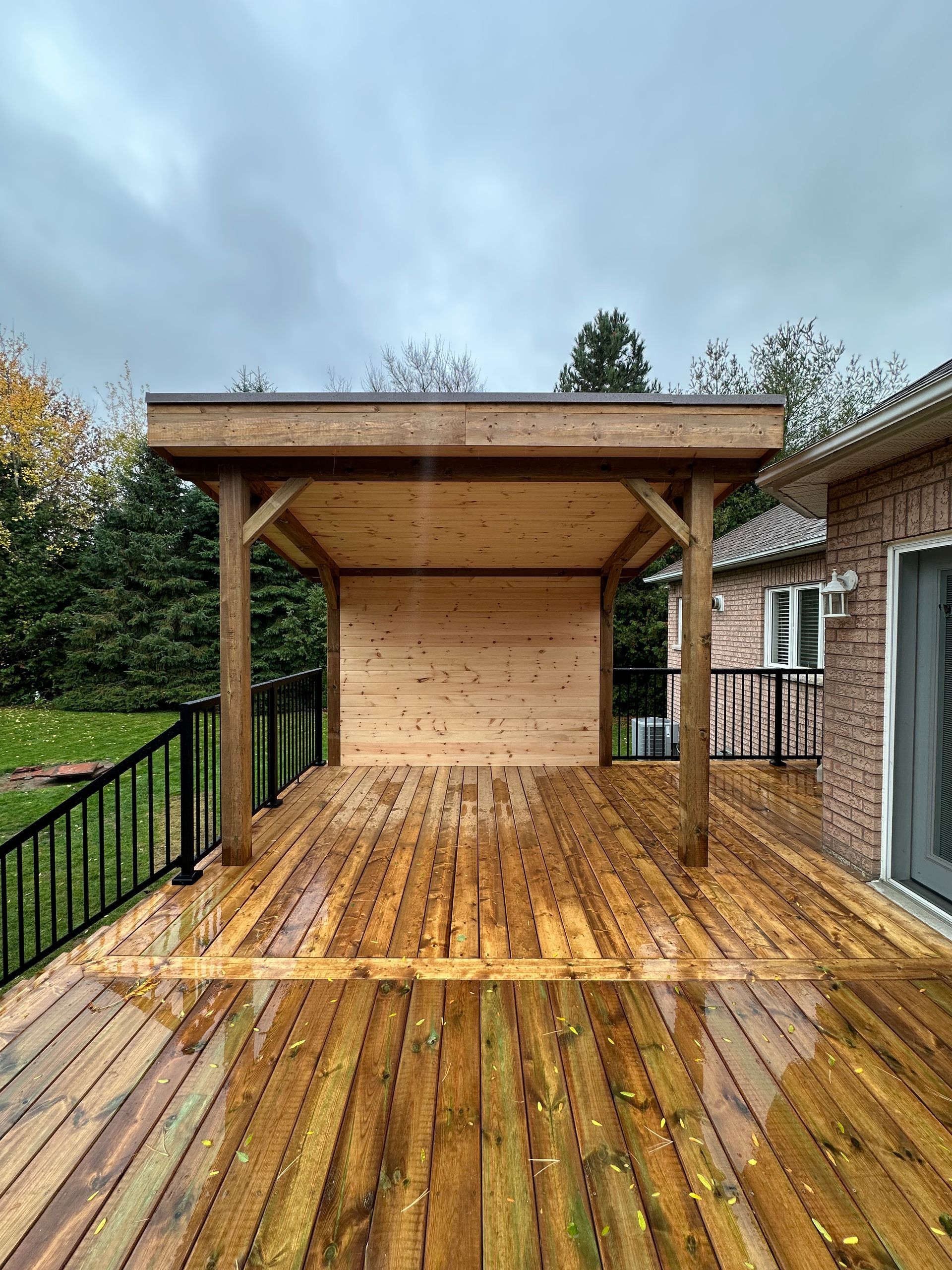 Wooden deck with a covered structure, black railing, and a brick house in the background on an overcast day.