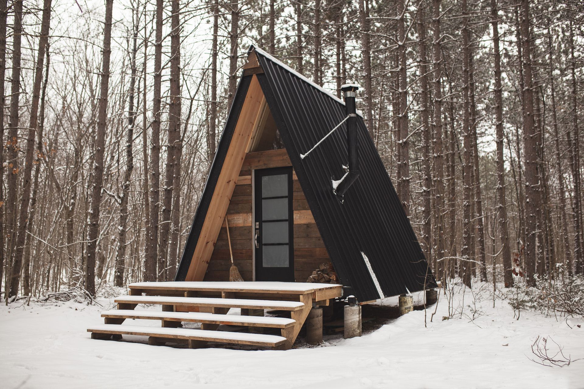 A-frame cabin in a snowy forest. Black roof and wooden steps, surrounded by bare trees.