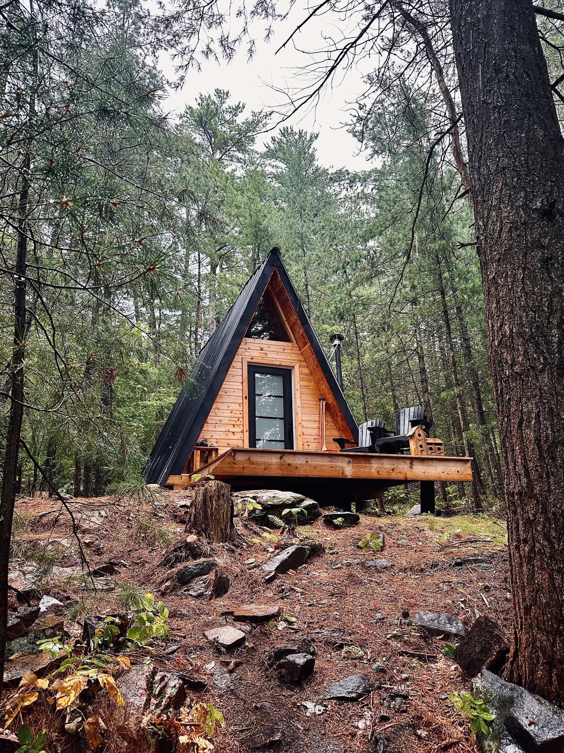 A-frame cabin nestled in a forest, wooden construction, black roof, deck, surrounded by trees and rocks.