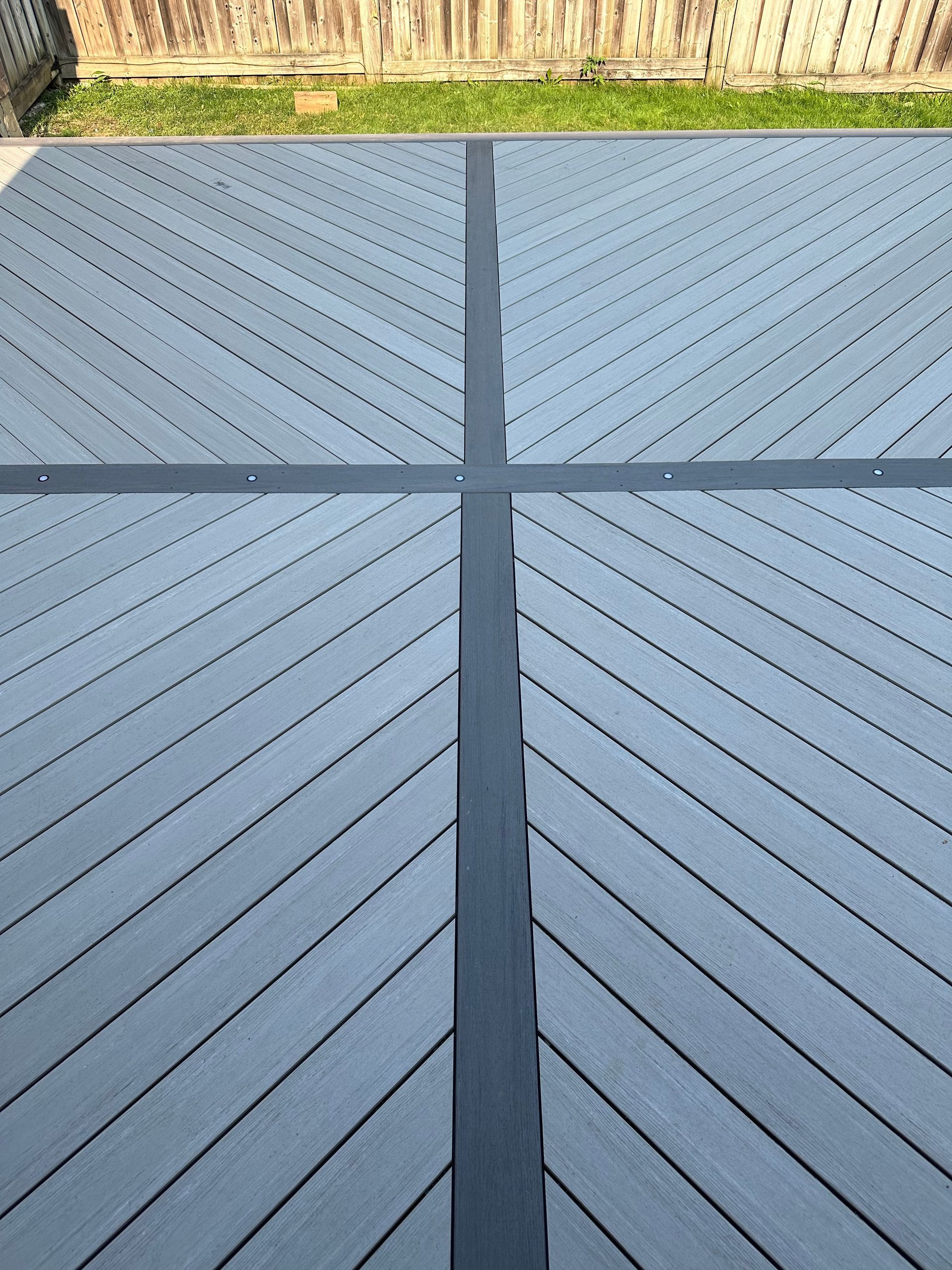Gray composite deck with angled planks, a dark cross pattern, and a wooden fence.