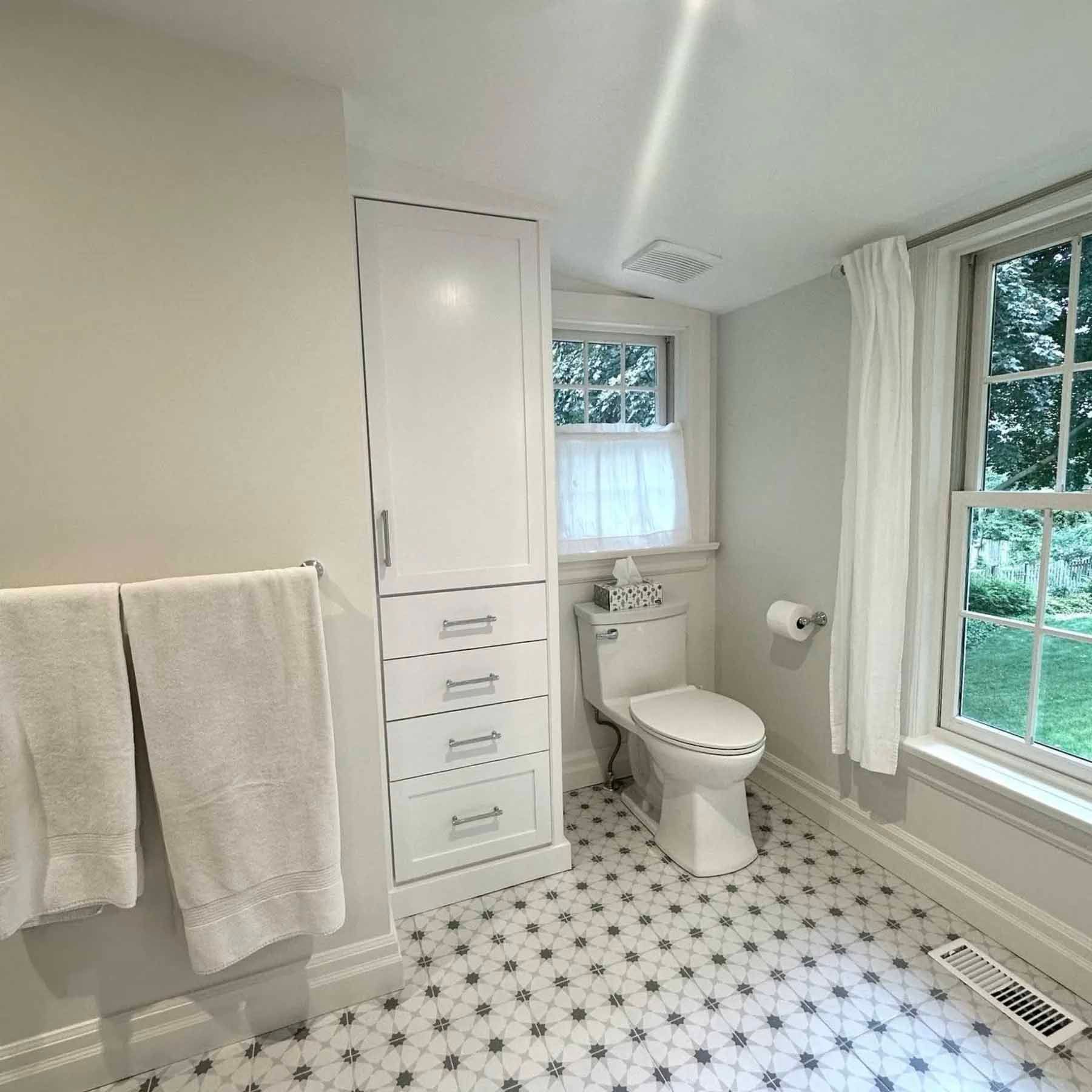 Bathroom with white cabinetry, toilet, patterned floor, and two windows with sheer curtains.