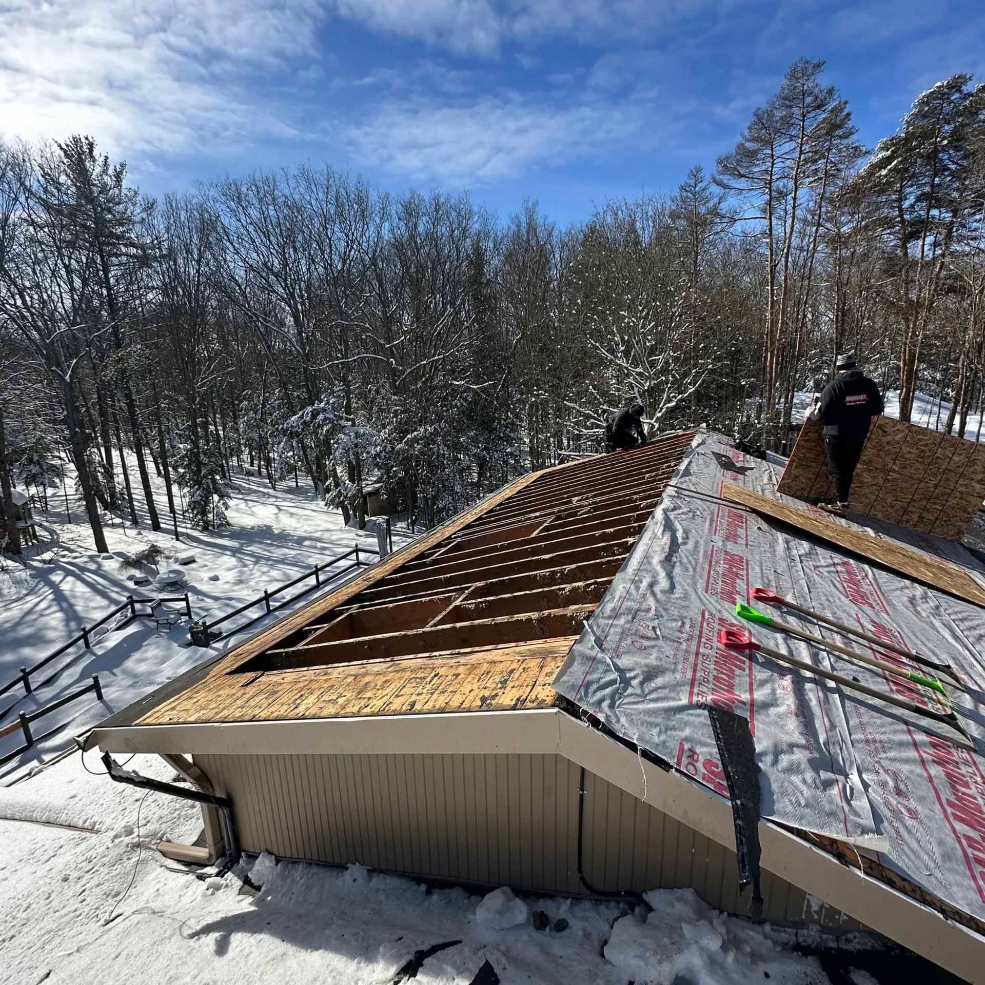 Roofers on a snowy roof installing shingles; trees and a blue sky in the background.