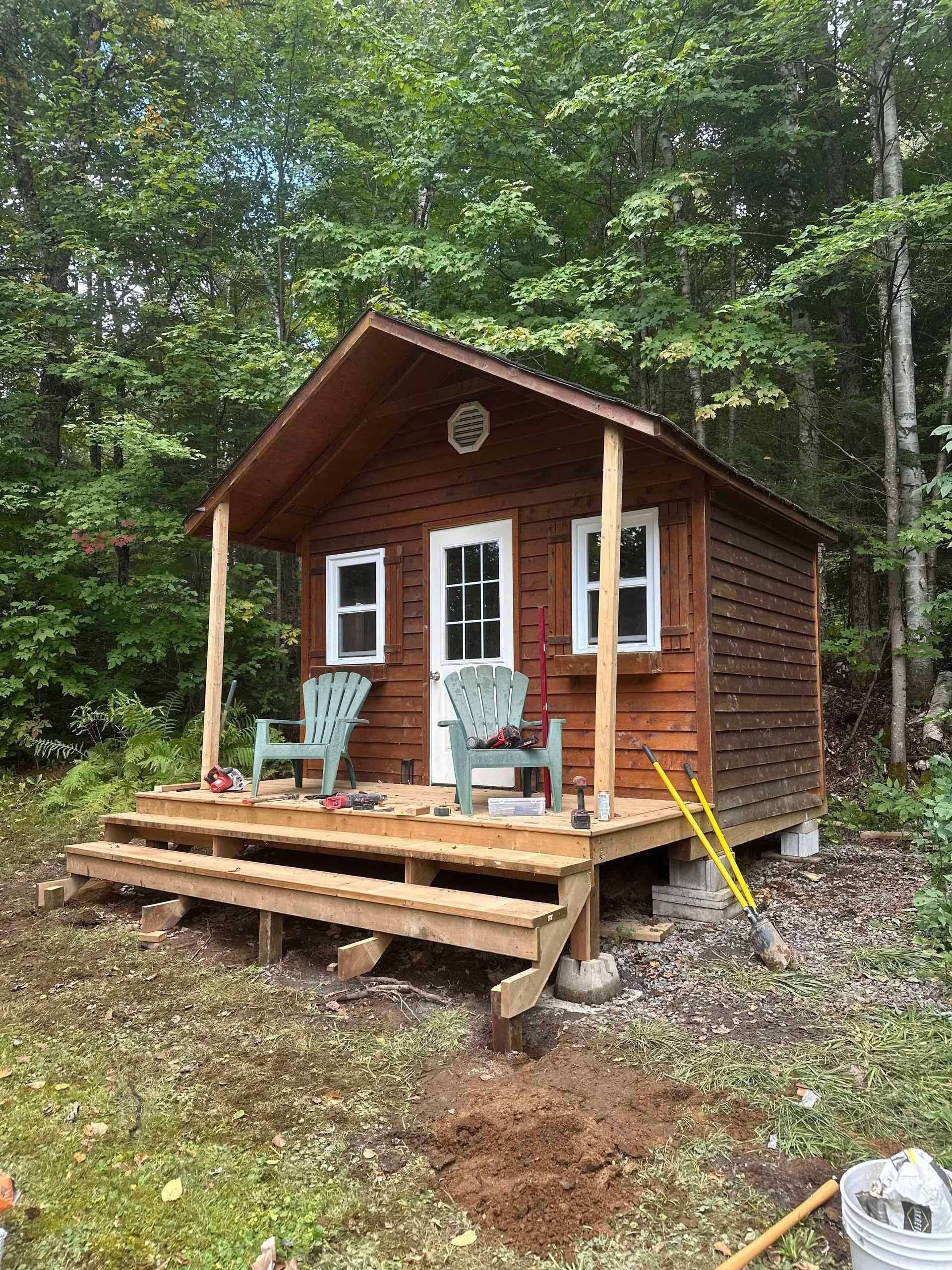 Small brown cabin with porch and two green chairs, set in a wooded area.