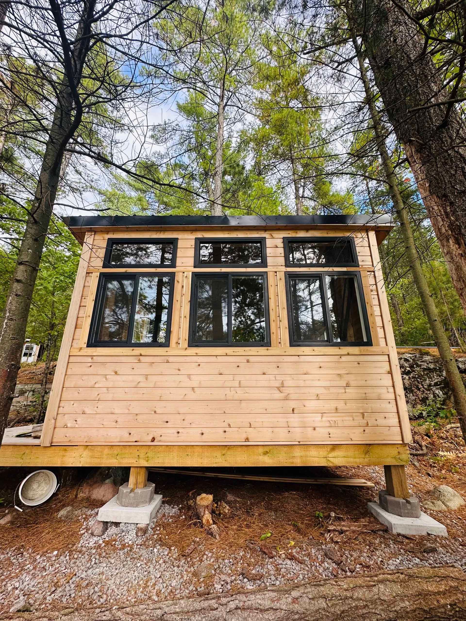 Wooden cabin with black-framed windows in a forest setting, elevated on concrete blocks.