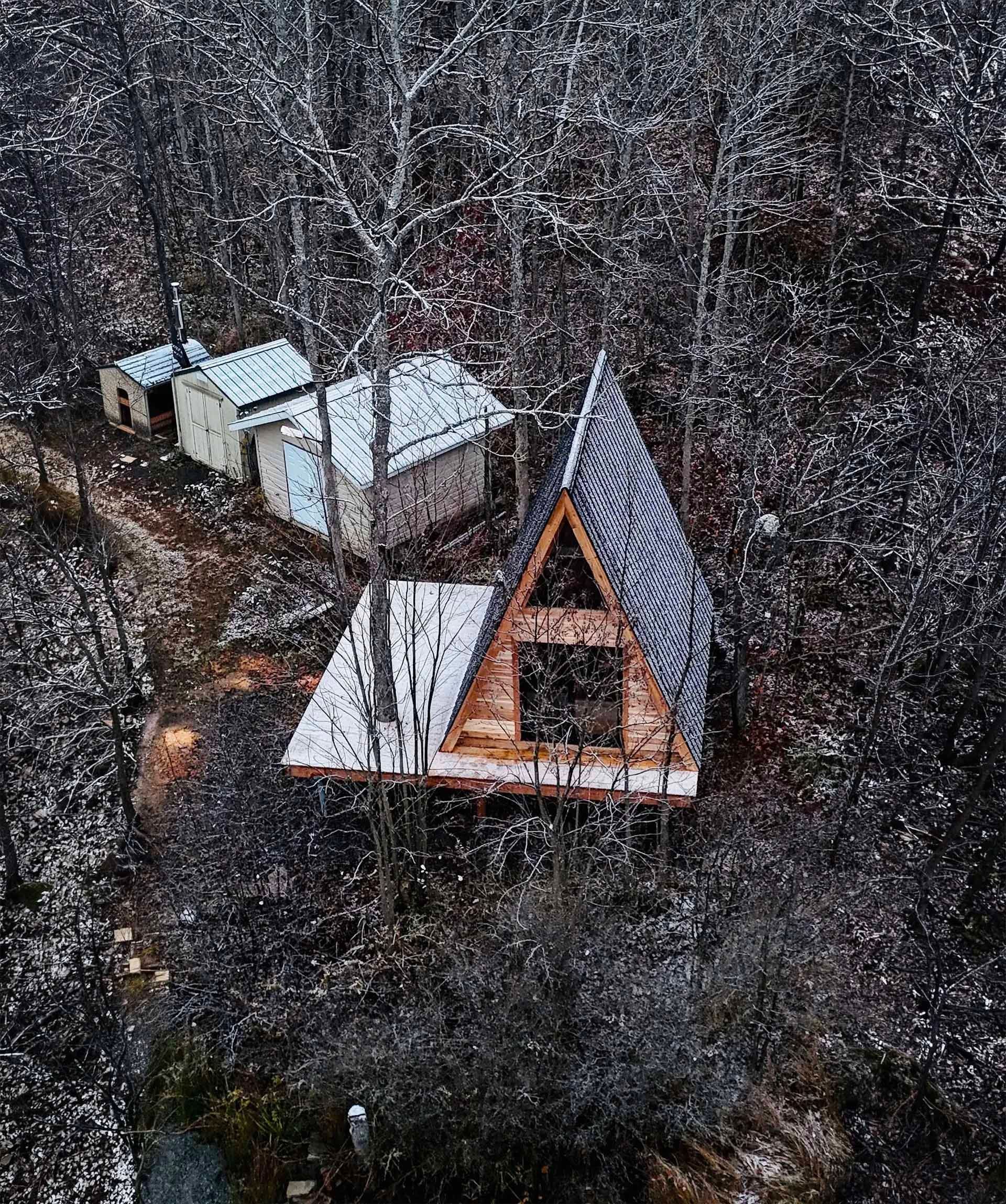 Wooden A-frame cabin and outbuildings in a snowy forest setting.