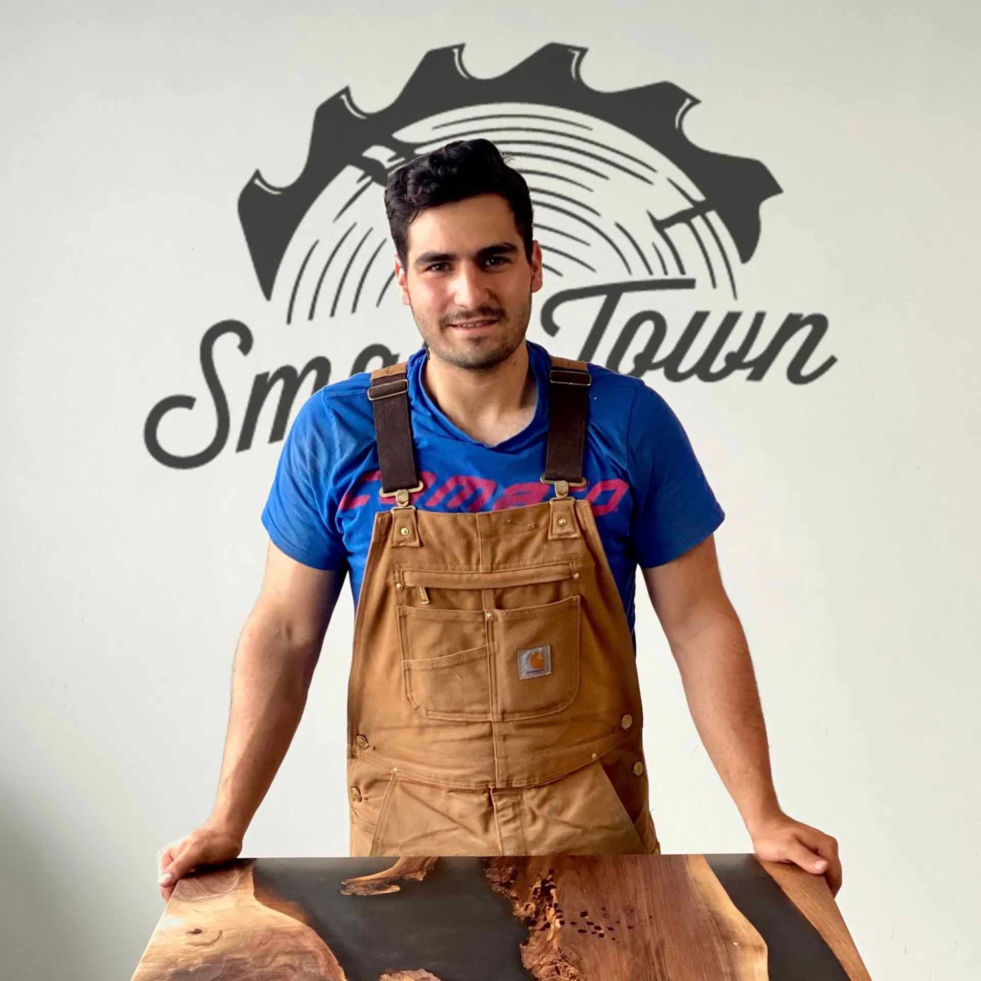 Man in brown overalls, standing in front of a wood slab, with the logo 