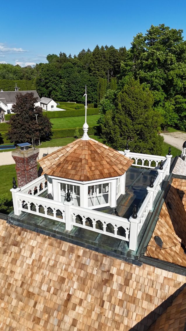 Octagonal gazebo with brown shingle roof, white trim, on rooftop with surrounding green landscape.