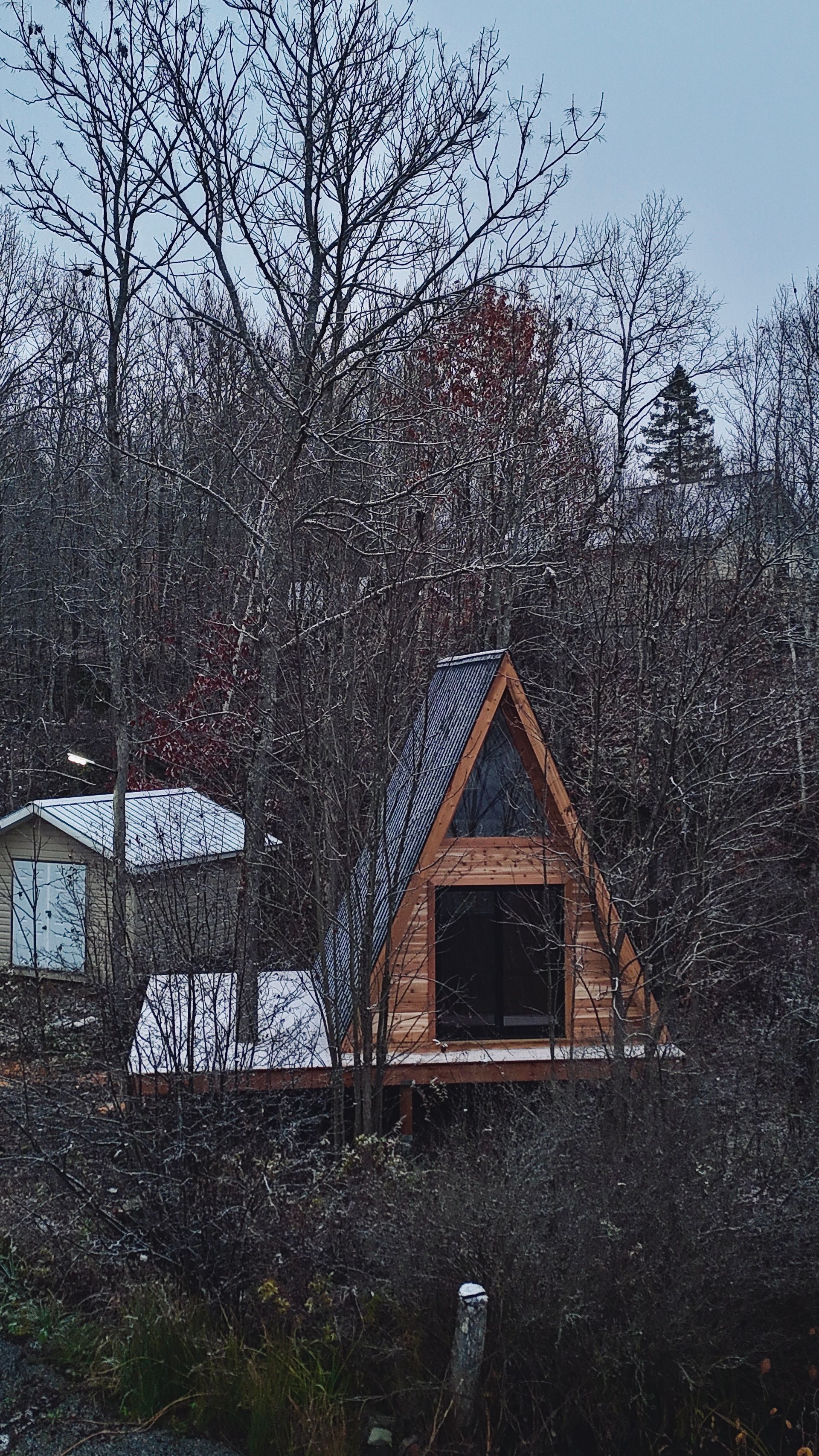A-frame cabin nestled in snowy woods with small shed visible.
