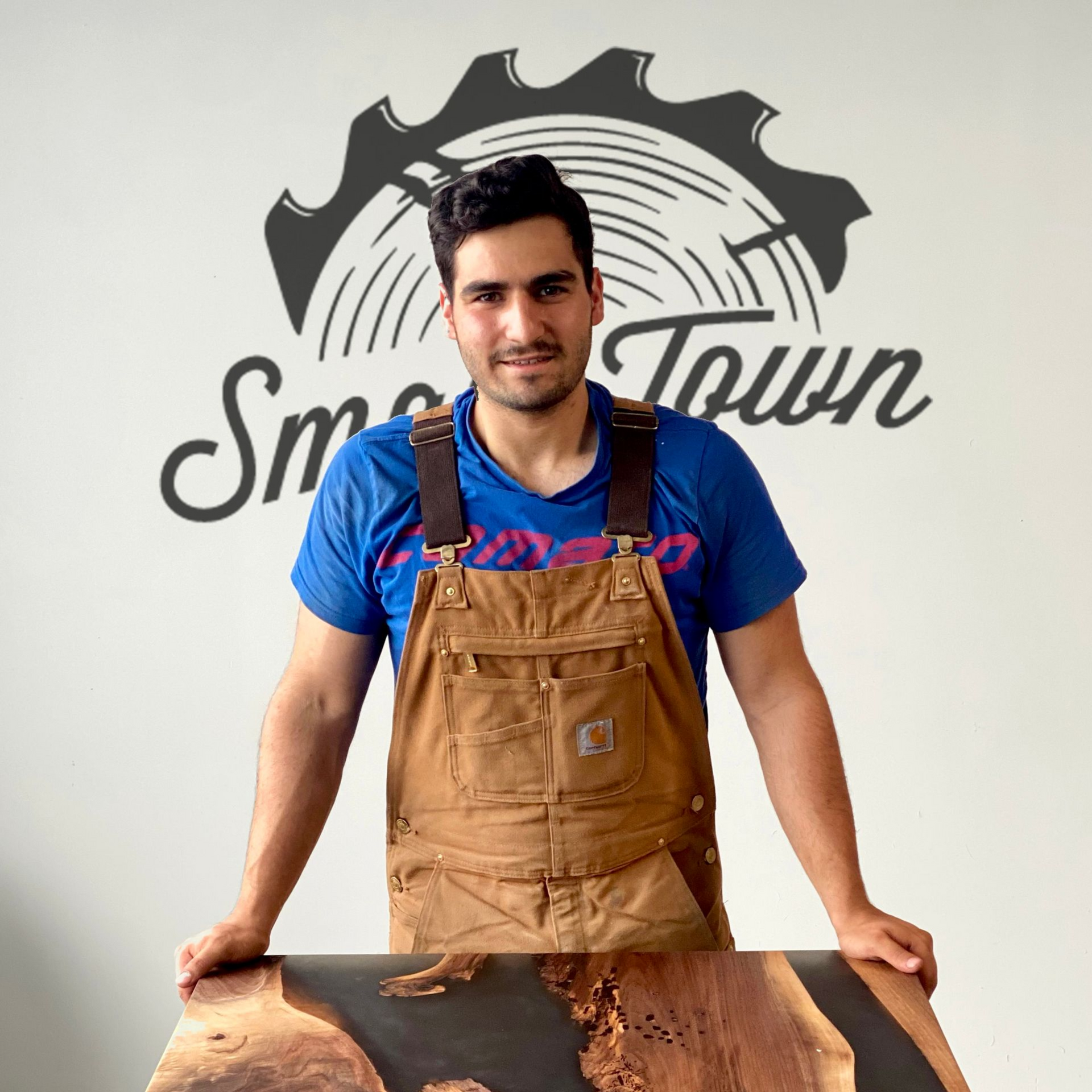 Man in brown overalls, standing in front of a wood slab, with the logo 