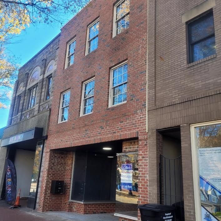 Three-story brick building between two other buildings; black storefront entrance on bottom.