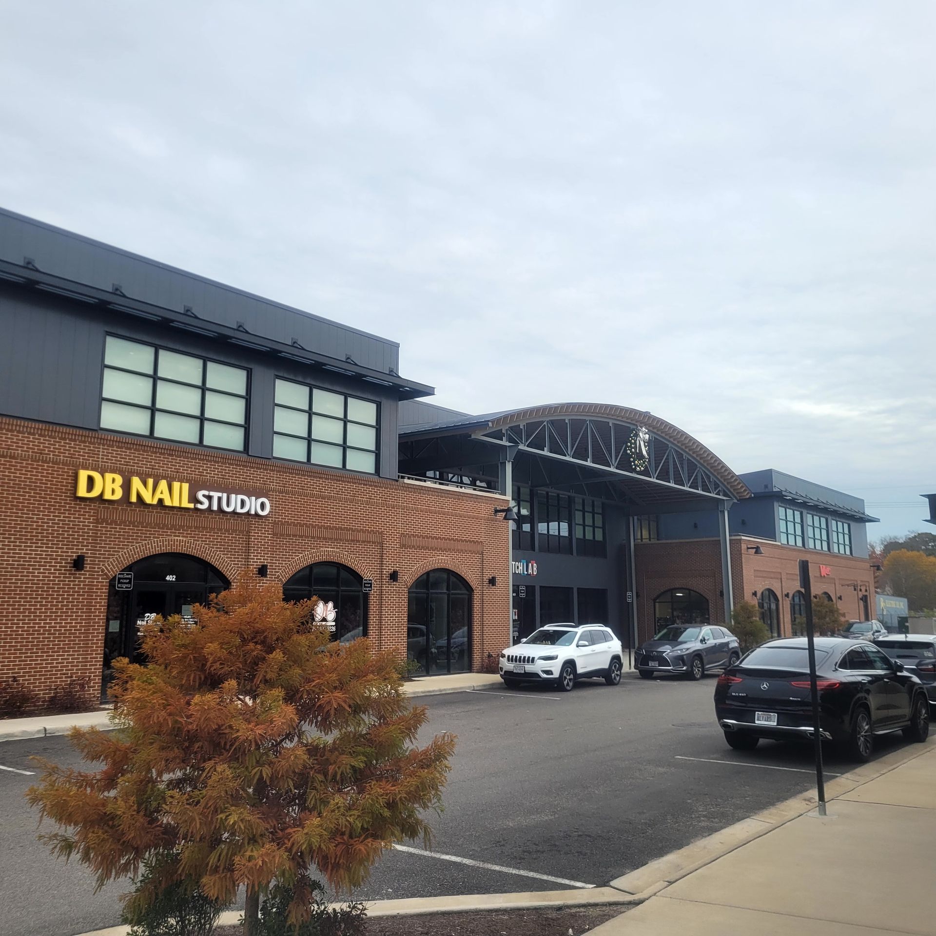 Exterior of a building with a nail salon sign, cars parked out front, and a tree with red leaves.