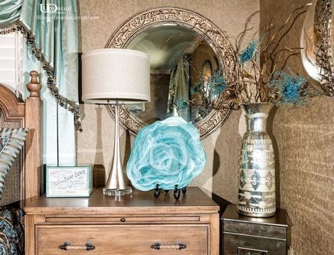 Nightstand with lamp, mirror, decor. Teal accents against neutral tones, wooden furniture.