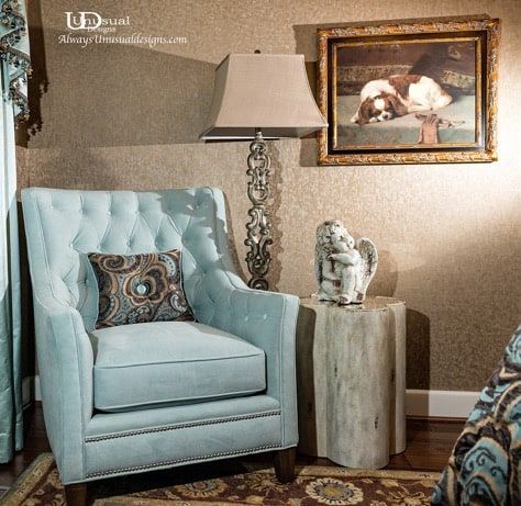 Light blue armchair with a patterned pillow, next to a wood side table and decorative lamp, with a painting of a dog on the wall.