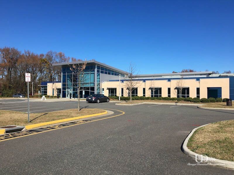 Modern building with glass entrance, blue accents, and parking lot on a sunny day.