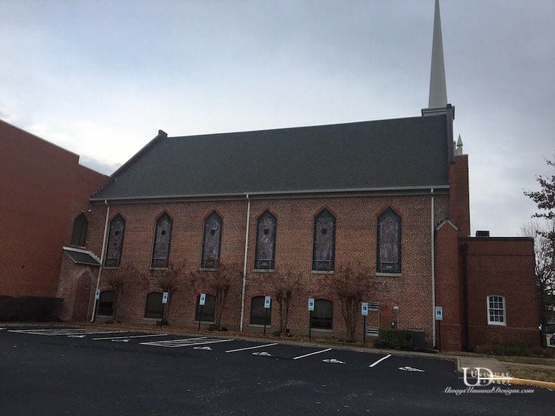 Brick church building with a steeple and stained glass windows, in a parking lot.