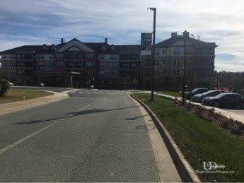 Asphalt road curves towards a large multi-story building under a blue sky. Cars parked to the right.
