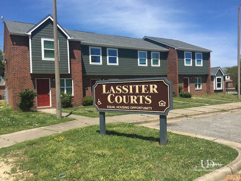 Lassiter Courts sign in front of multi-unit brick and green buildings with red doors and gray roofs.