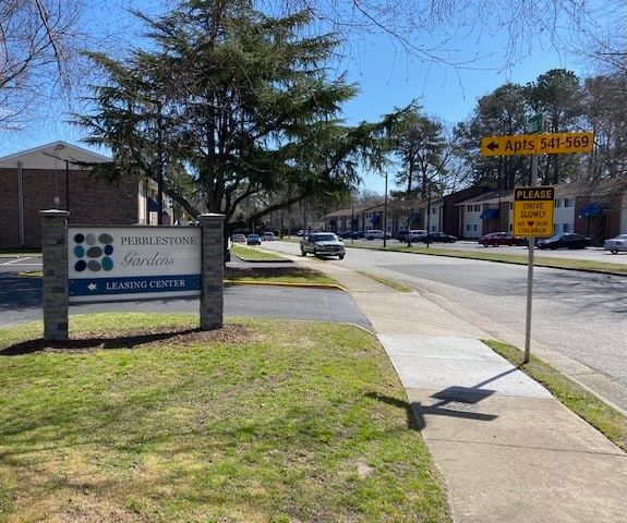 Entrance sign for Pebblebrook apartments, a street, and residential buildings under a blue sky.