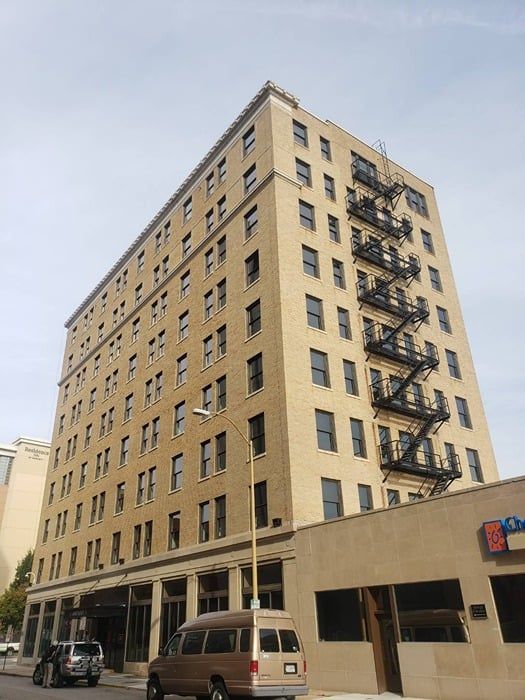 Tan brick multi-story building with fire escape, vehicle parked nearby.