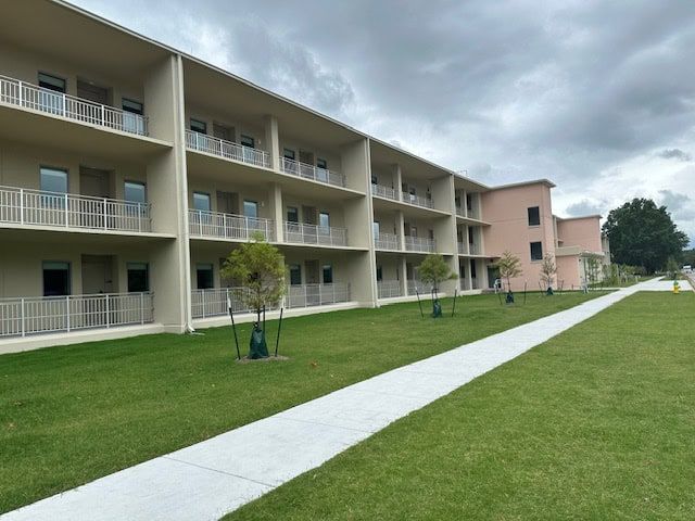Multi-story building with balconies, beige and pink exterior, sidewalk, green lawn, cloudy sky.