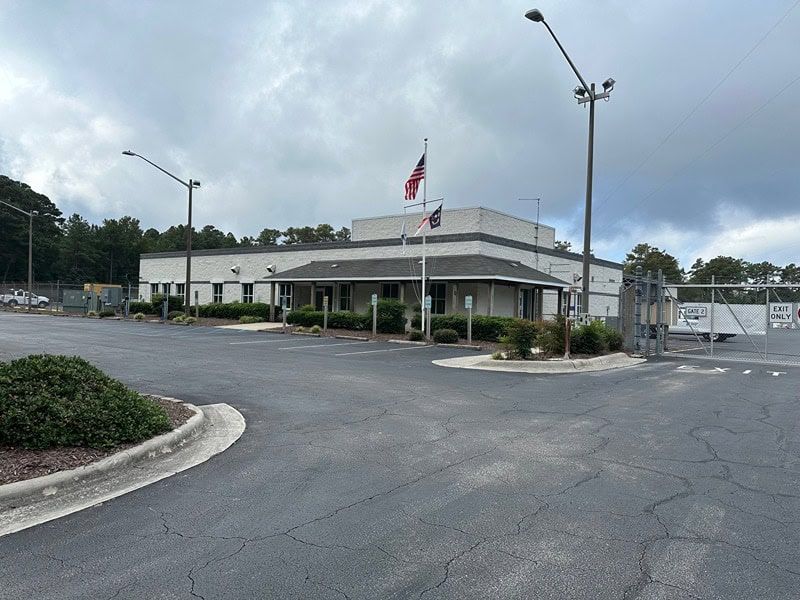 A low-slung building with an American flag flies in front. A gated entrance sits to the right, cloudy sky.