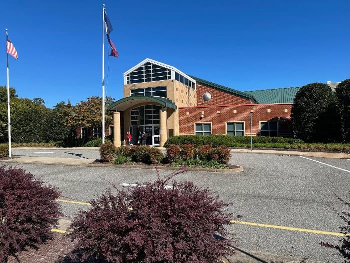 A brick building with flags in front, bushes, and a paved circular driveway on a sunny day.