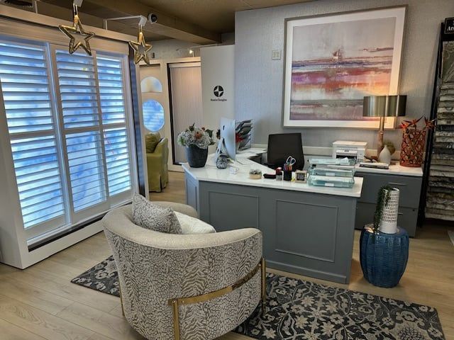 Office reception area with gray desk, patterned chair, art, and window shutters.