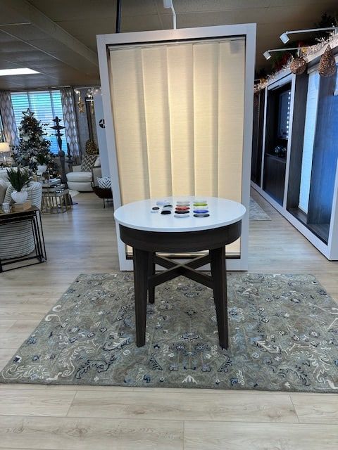 A table with samples sits on a rug in front of a vertical blind display. Interior design showroom setting.