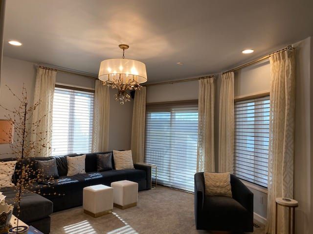 Living room with sofa, chair, ottoman, and sheer curtains. Gold chandelier hangs from ceiling.