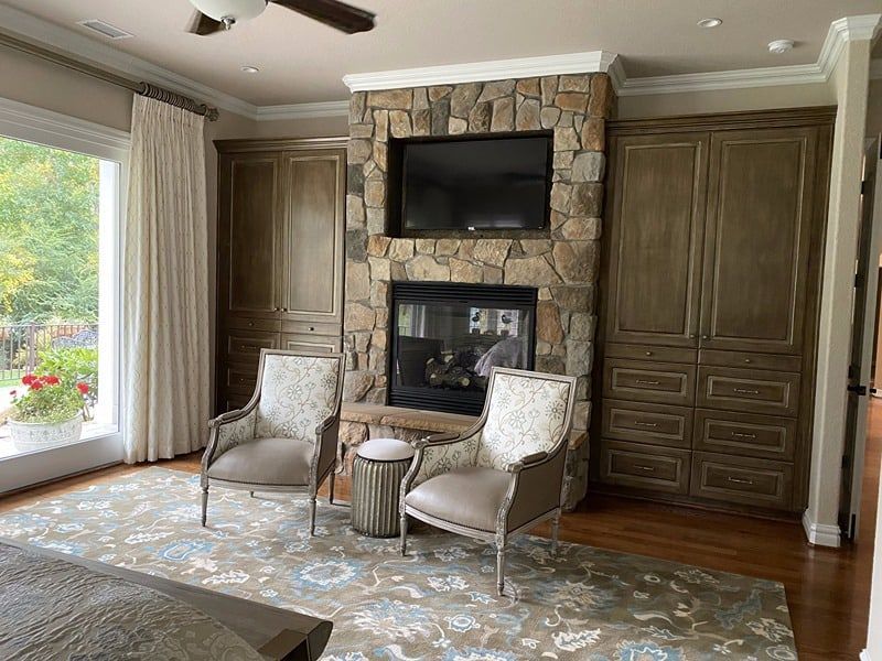 Cozy bedroom with stone fireplace, TV, built-in cabinets, two armchairs, and a patterned rug.