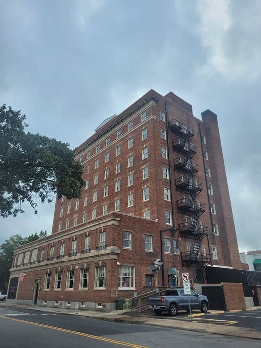Brick multi-story building with fire escape, cloudy sky, parked vehicles, and street.