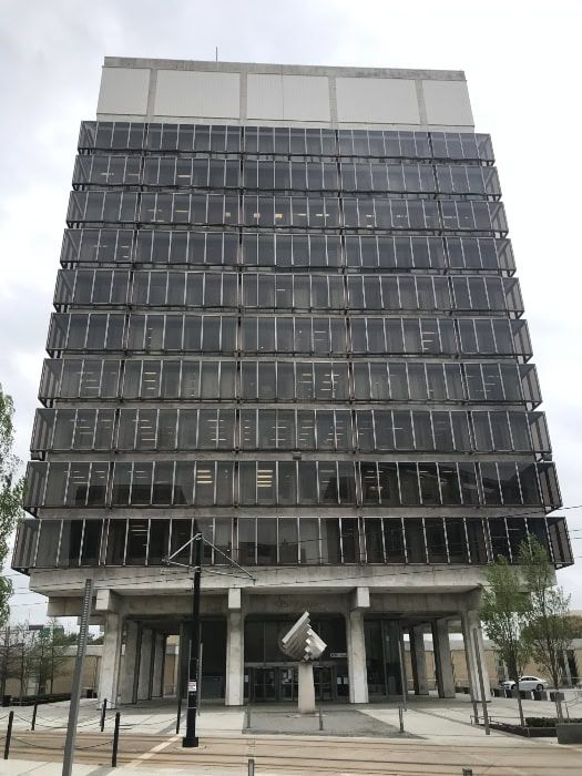 Modern multi-story building with a square facade and numerous windows, under a cloudy sky.