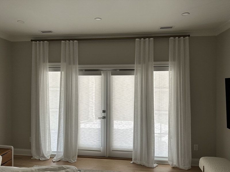 Sheer white curtains hang over a set of glass doors and windows. Brown curtain rod. Neutral-colored walls.