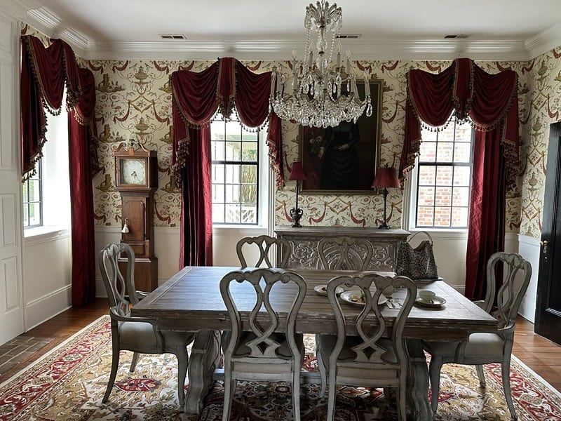 Elegant dining room with red drapes, chandelier, and a long wooden table set for six.