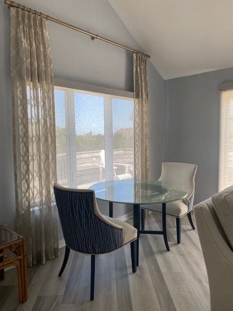 Dining area with round glass table, chairs, and sheer curtains. Gray walls and light wood floors.