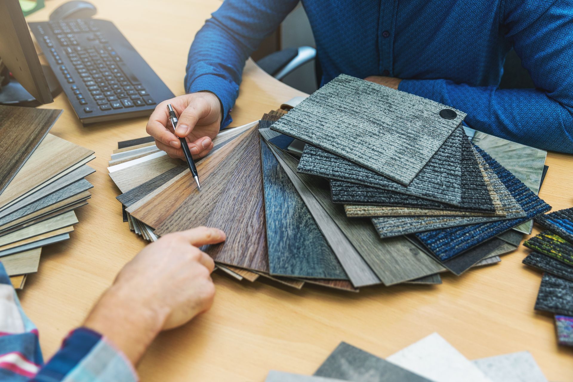 Two people examine flooring samples at a desk, one pointing with a pen.