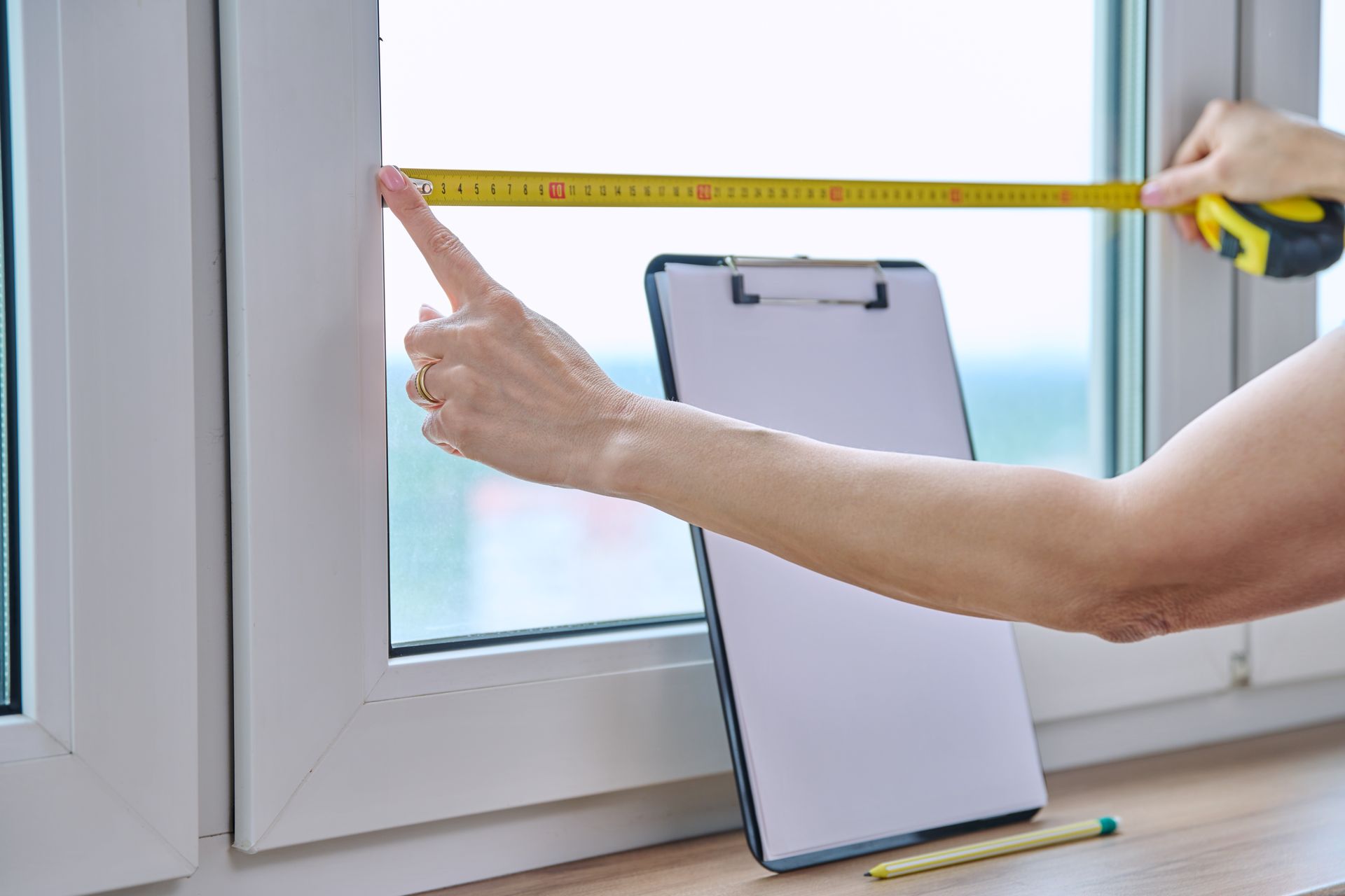 Person measuring a window with a tape measure; clipboard and pencil on the sill.