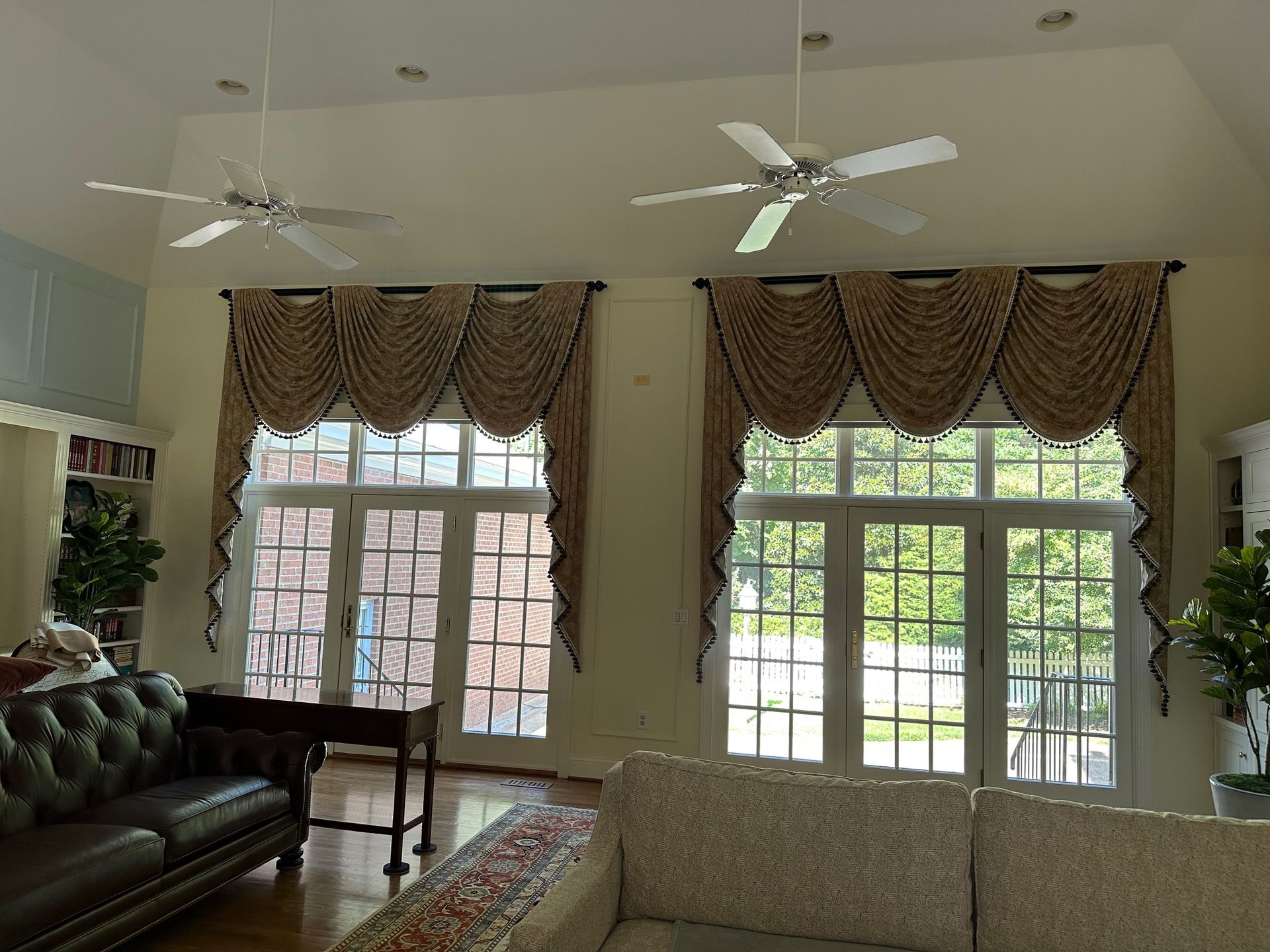 Two windows with ornate gold and black drapery, flanked by white ceiling fans, in a living room.