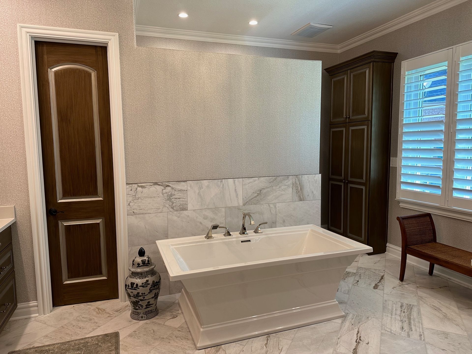 Bathroom with white tub, marble tile, brown door, and shutters.