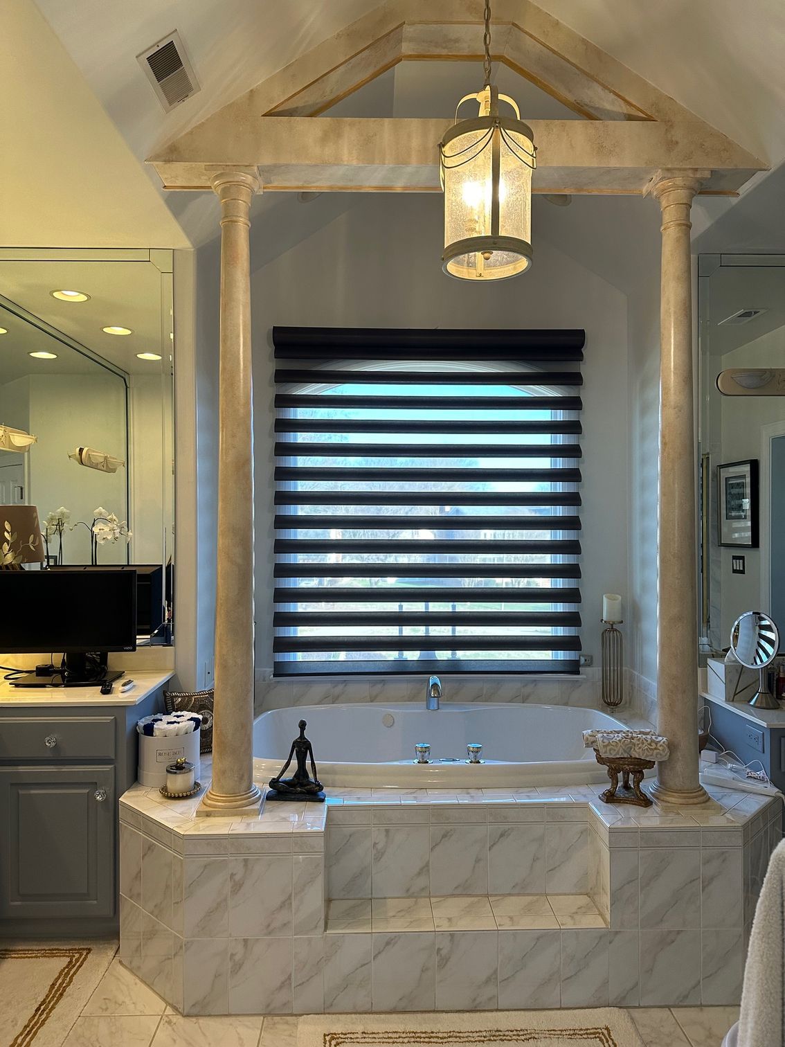 Bathroom with built-in jacuzzi, ornate columns, and zebra-style window shade. Beige and white tile, pendant light.