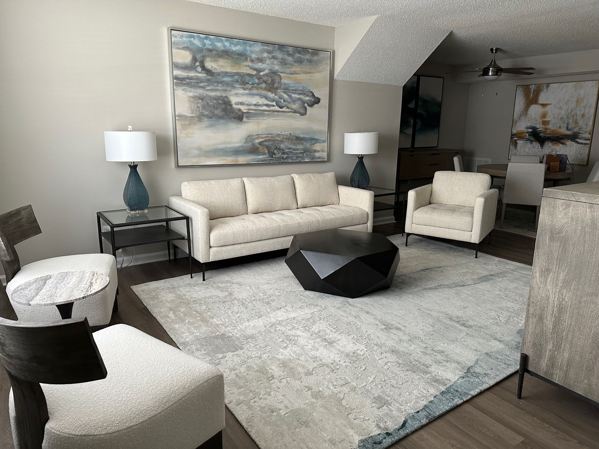 Living room with beige sofa, chairs, art, black coffee table, lamps, and rug.