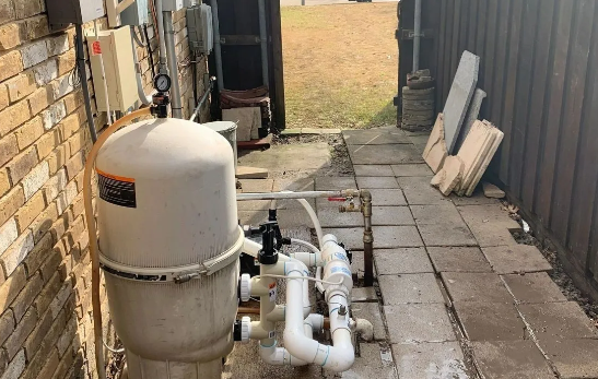 Pool equipment, including a white filter tank, in a narrow outdoor space with brick and wooden fencing.