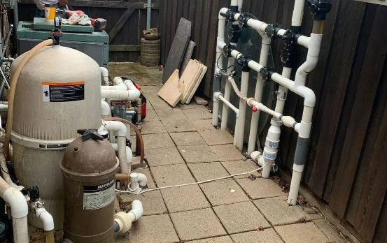 Pool equipment: filter tanks, plumbing, and pipes against a wooden fence in an outdoor area.