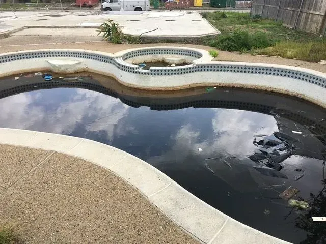 Dirty, abandoned swimming pool with murky water reflecting cloudy sky.