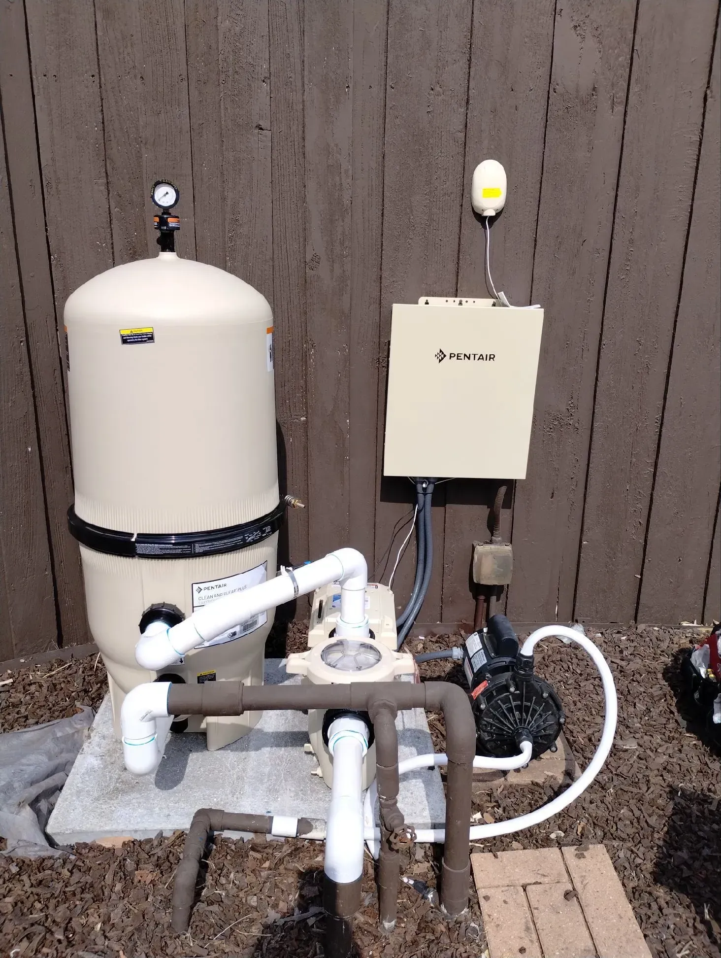 Water well system with a beige tank, pump, and electrical box against a brown wooden fence.