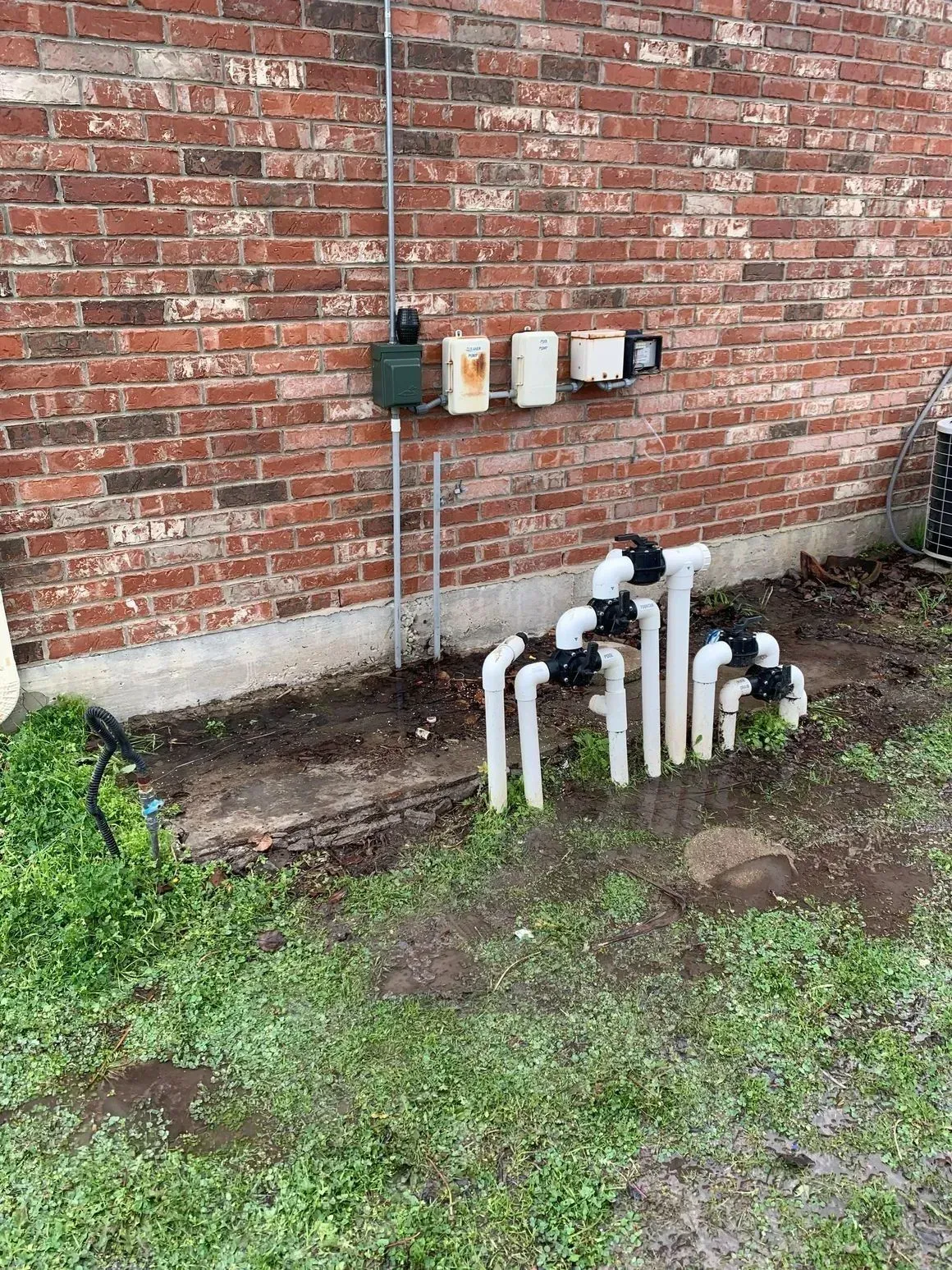 Brick wall with electrical boxes, PVC pipes, and valves in a grassy area.